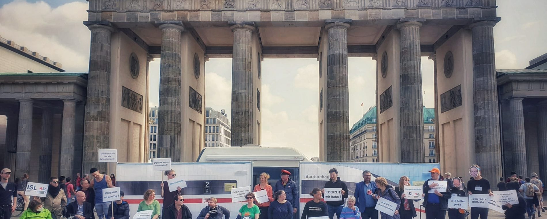 ISL e.V. Bahnaktion zum Protesttag 5. Mai zum Thema Diskriminierung beim Bahnfahren. Eine Gruppe von Menschen mit Plakaten steht vor einem aufgebauten ICE. Im Hintergrund das Brandenburger Tor.